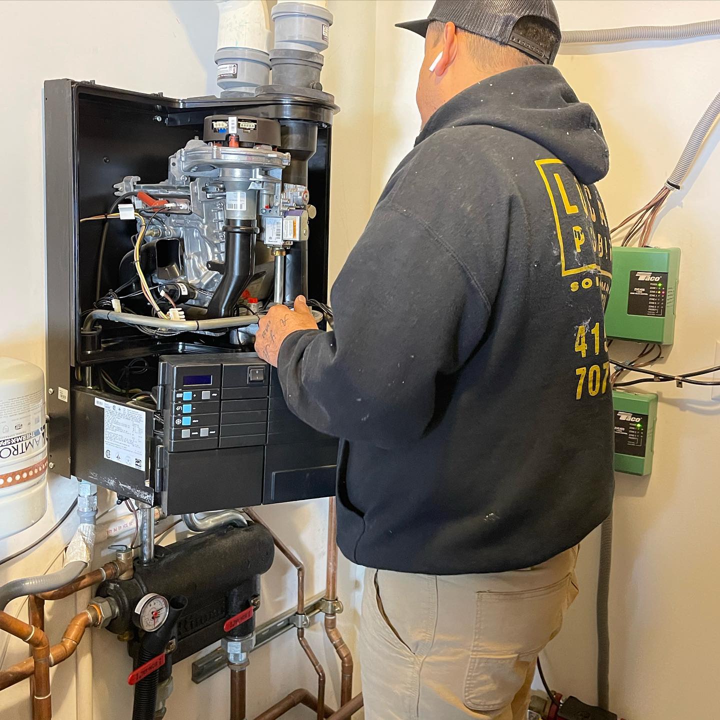 Person in a black hoodie inspecting a wall-mounted heating system with copper pipes.