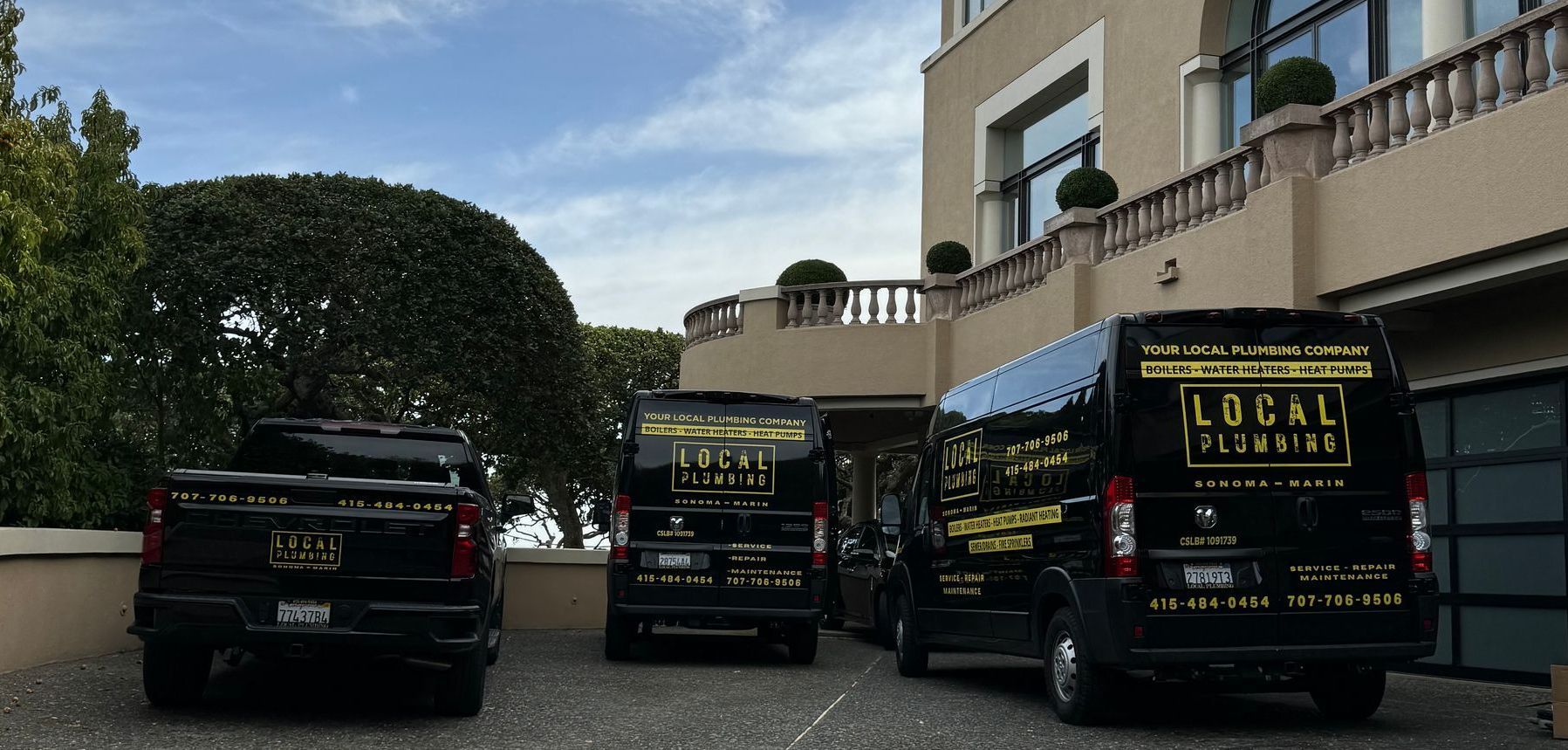 Three black plumbing vans and a truck parked near a large building with a green hedge.