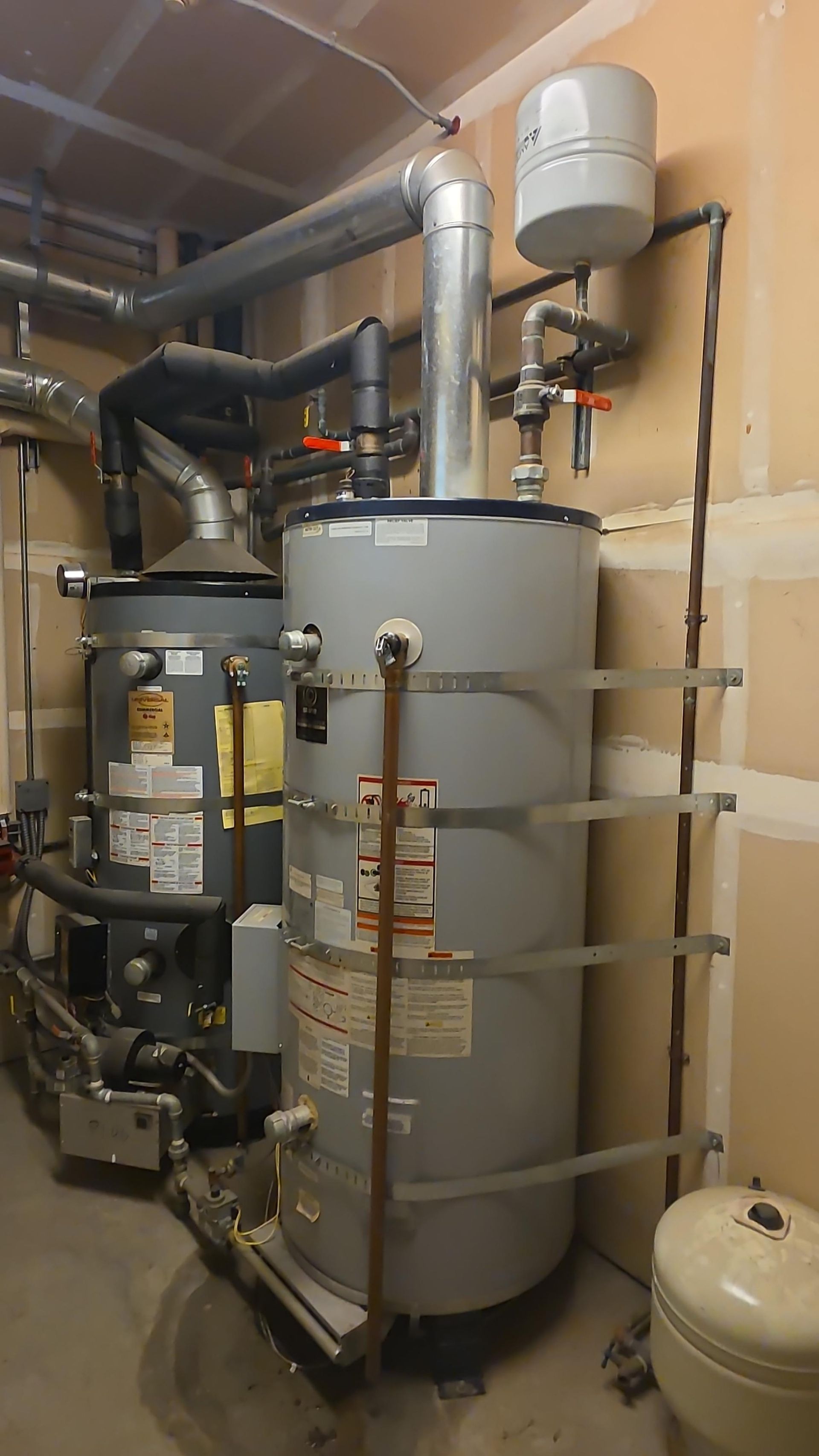 Two water heaters with pipes and venting against a beige wall in a basement.