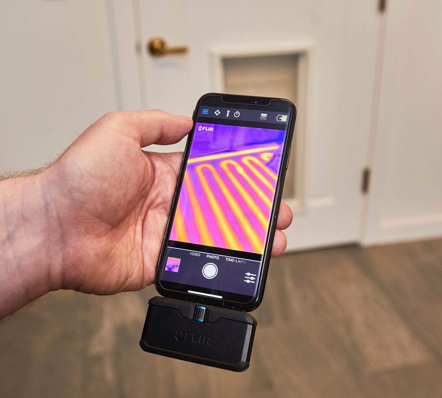 A hand holds a smartphone displaying a thermal image of a heated floor. The phone is attached to a black thermal camera.