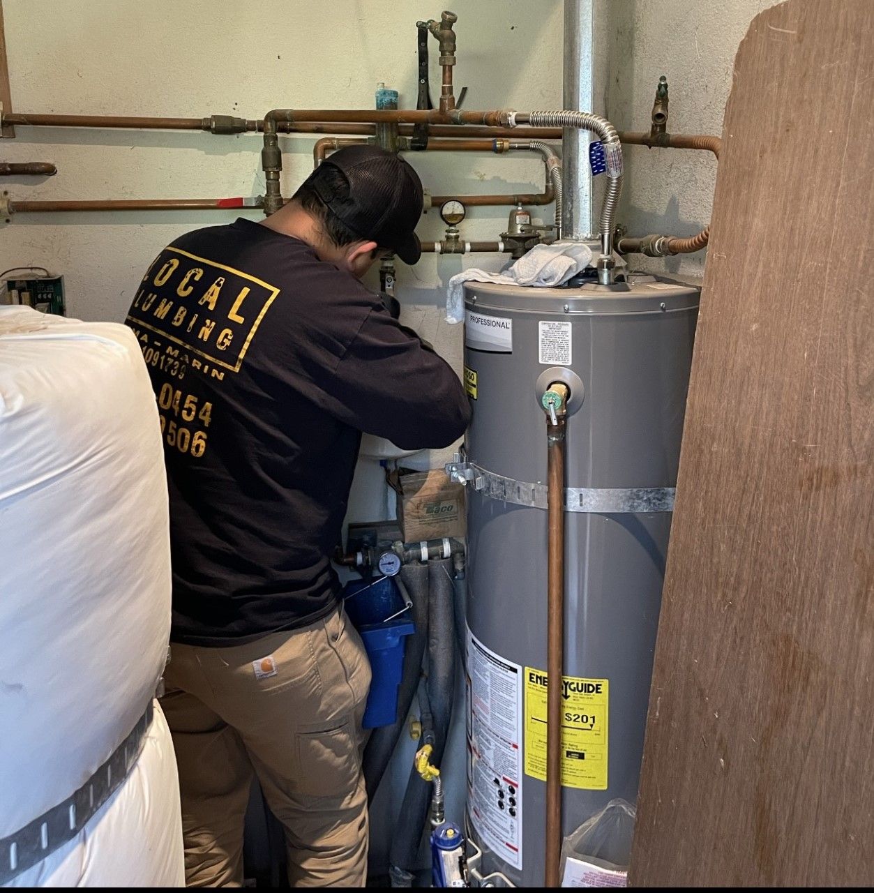 Plumber in black shirt and cap works on a water heater in a utility room. Copper pipes and insulation are visible.