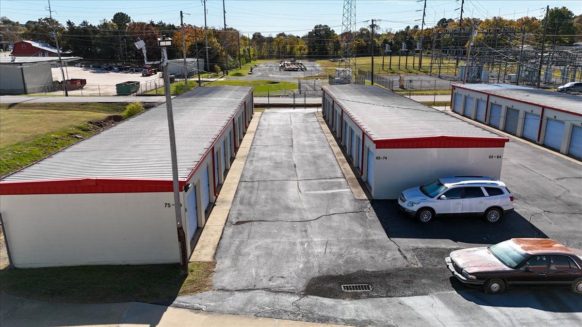 Exterior view of storage units, two rows facing each other with asphalt drive. White SUVs and sedan parked in front.