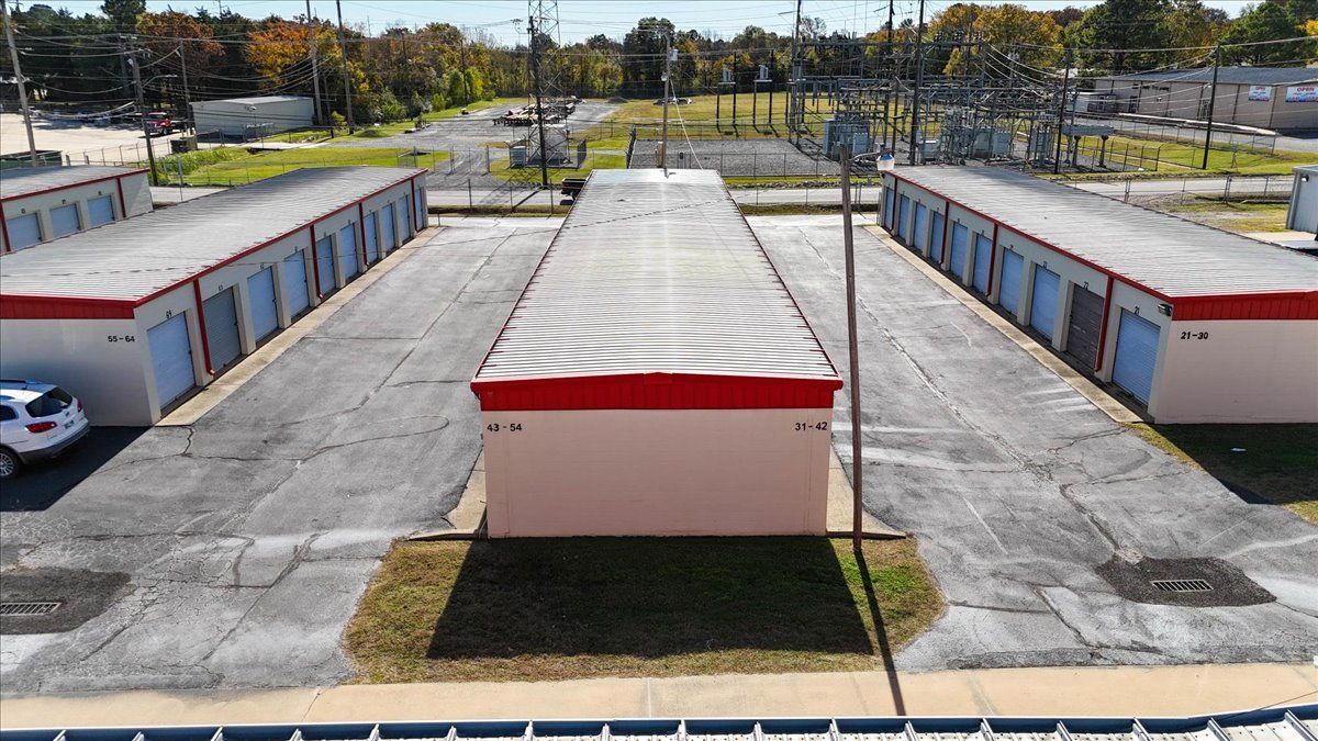 Overhead view of a storage facility with three rows of units and a large central building. Red roof accents.