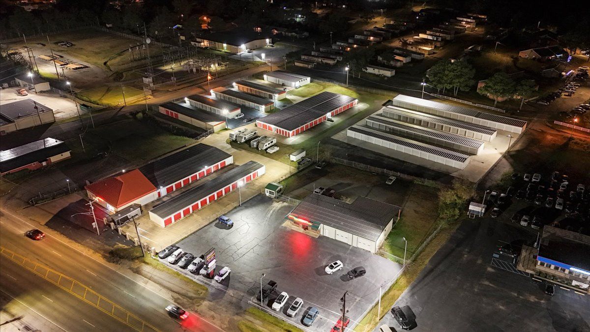 Night aerial view of a brightly lit commercial area with buildings, parking, and roadways.
