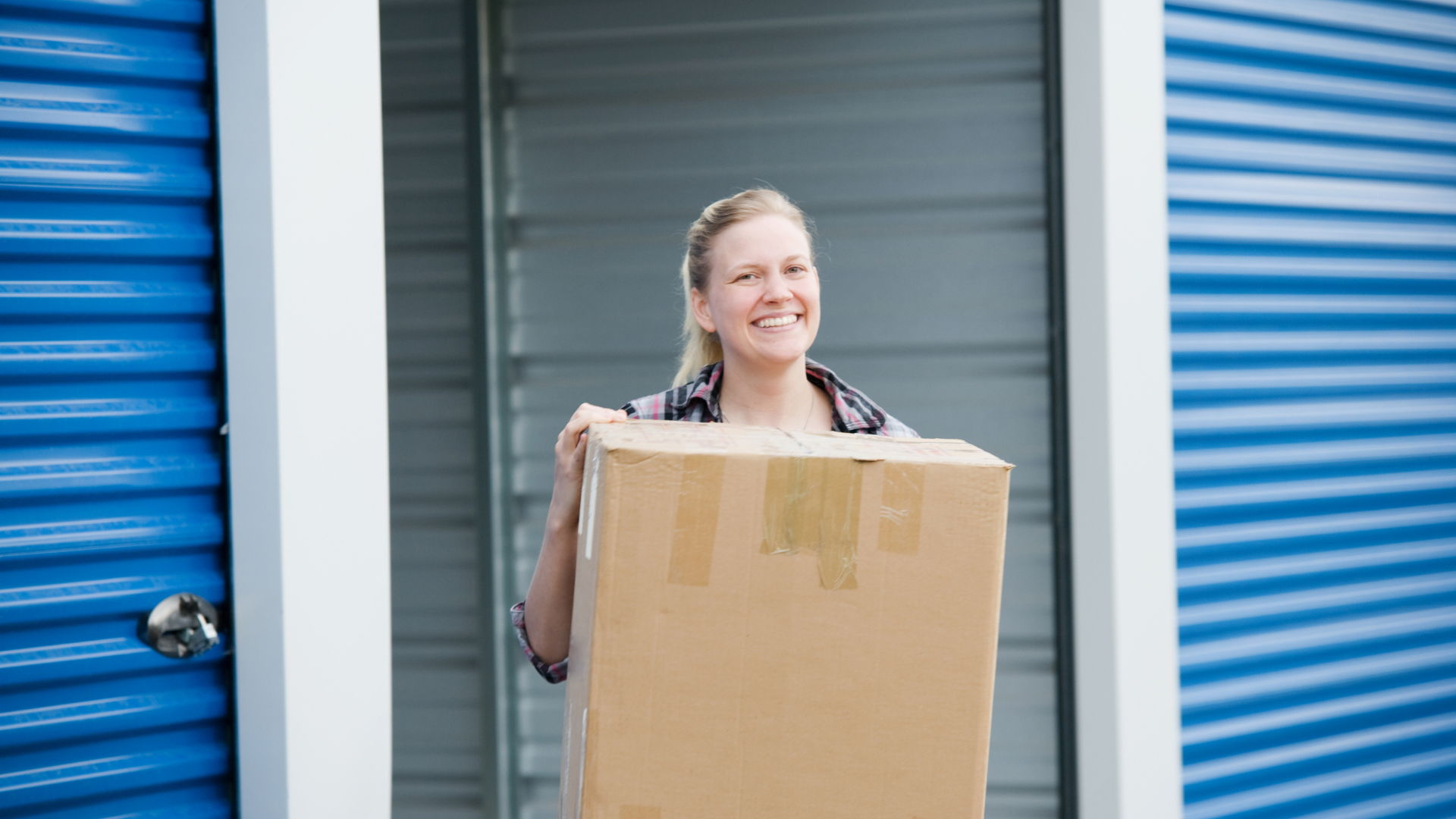 Woman carrying a cardboard box, smiling, exiting a blue storage unit.