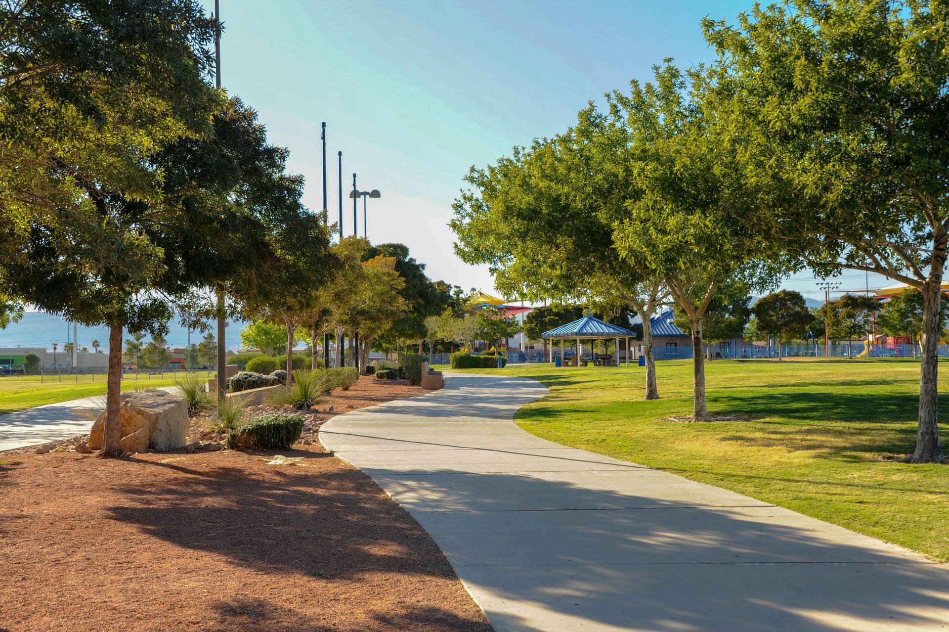 A paved walking path winds through a park with green grass, trees, and a small shaded pavilion under a clear sky.