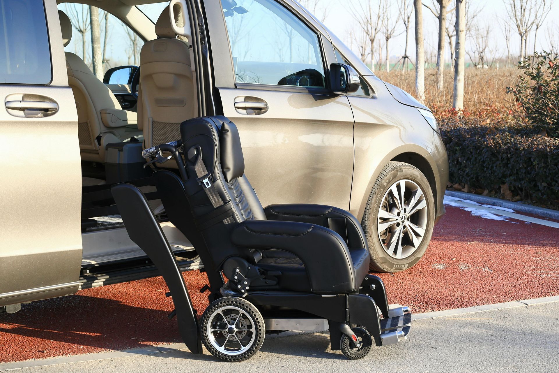A black power wheelchair with an extended lifting mechanism parked outside a beige van on a paved outdoor lot.