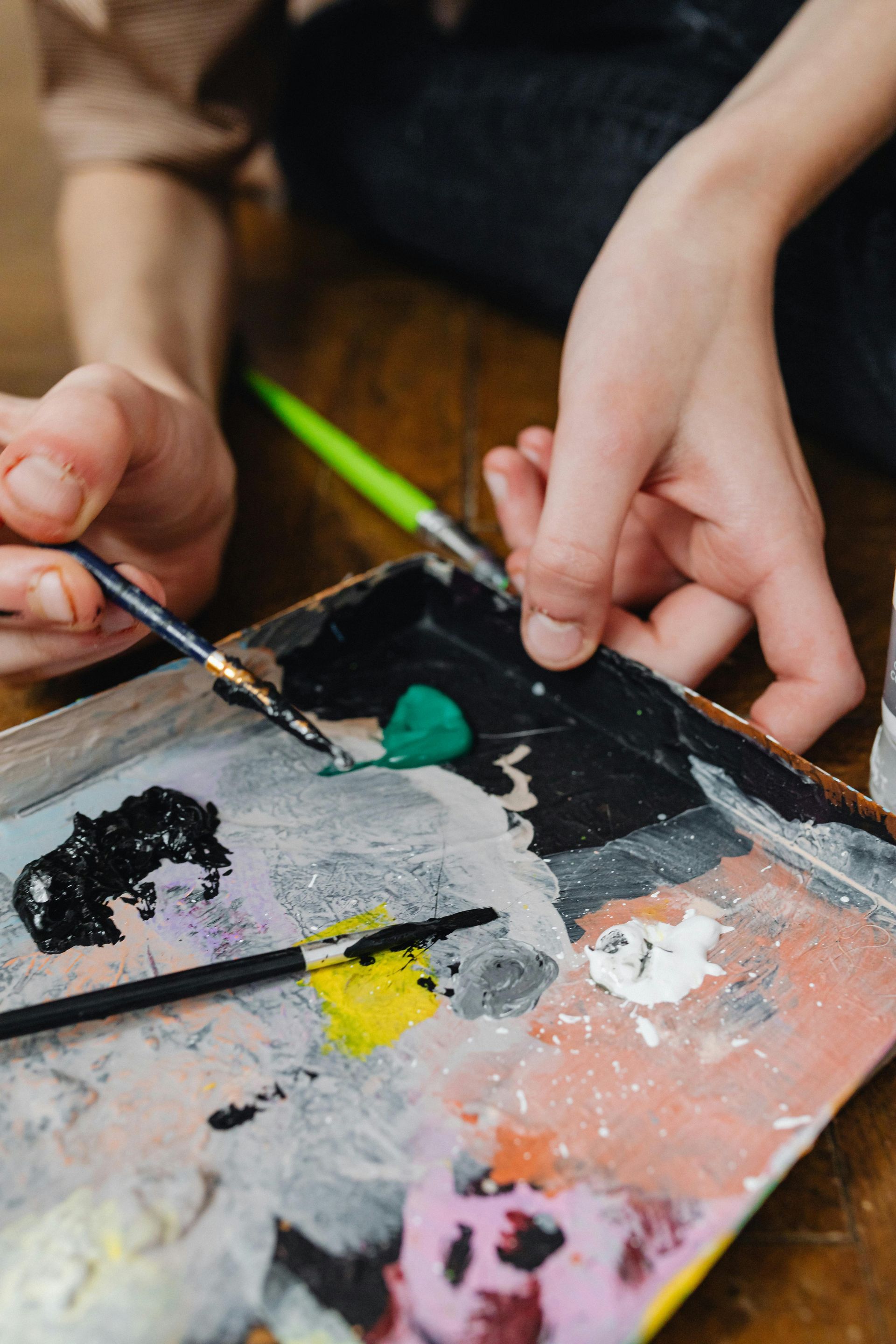Hands holding brushes, mixing colorful paints on a palette over a wooden floor.