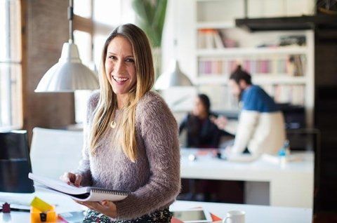 una persona sorridente tiene in mano un quaderno, mentre sullo sfondo si vedono due colleghi.