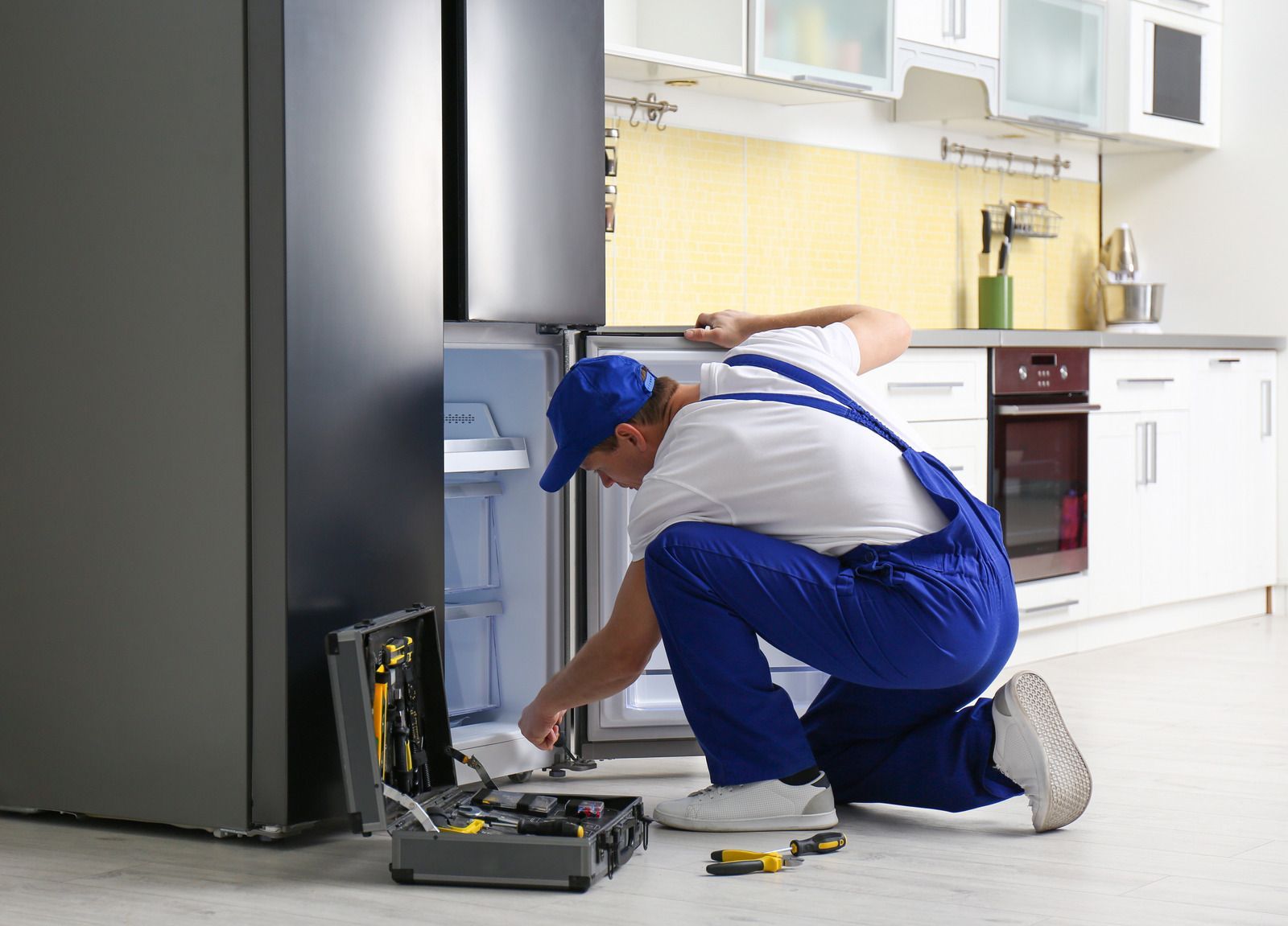 A repairman in blue overalls fixes a refrigerator in a kitchen, kneeling with a toolbox.