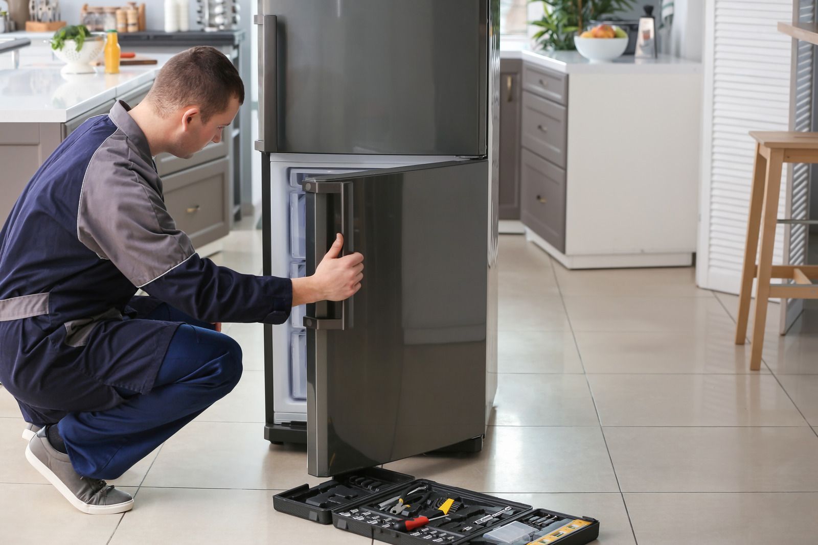 A person in work clothes inspecting a refrigerator door in a kitchen, tool kit on the floor.