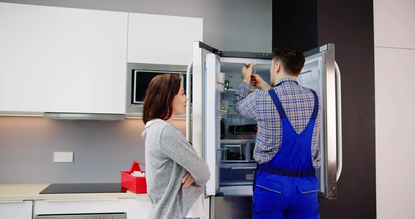 Woman watches a repairman working on a refrigerator in a modern kitchen.
