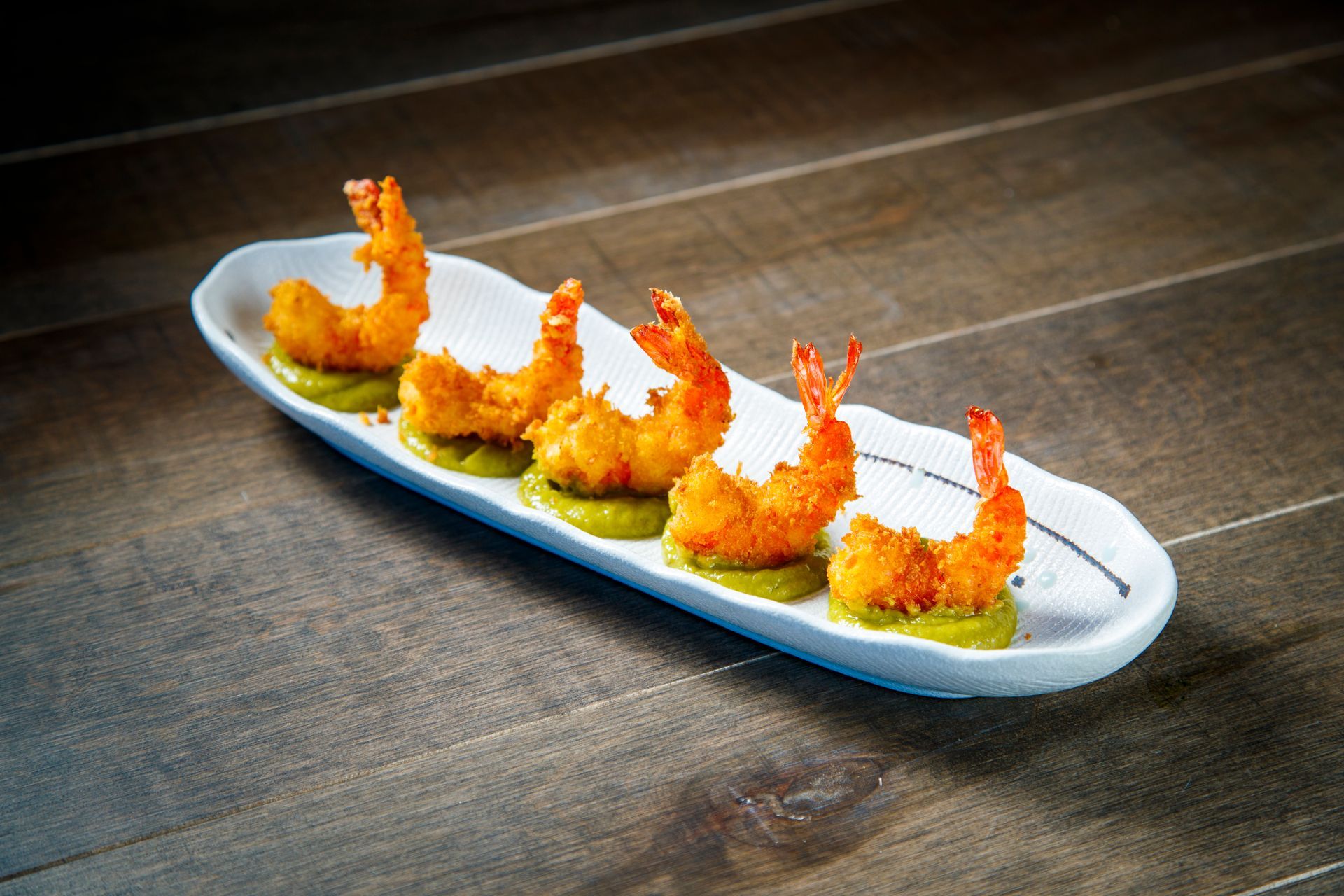 Five golden fried shrimp on a long white platter with green garnish on a dark table