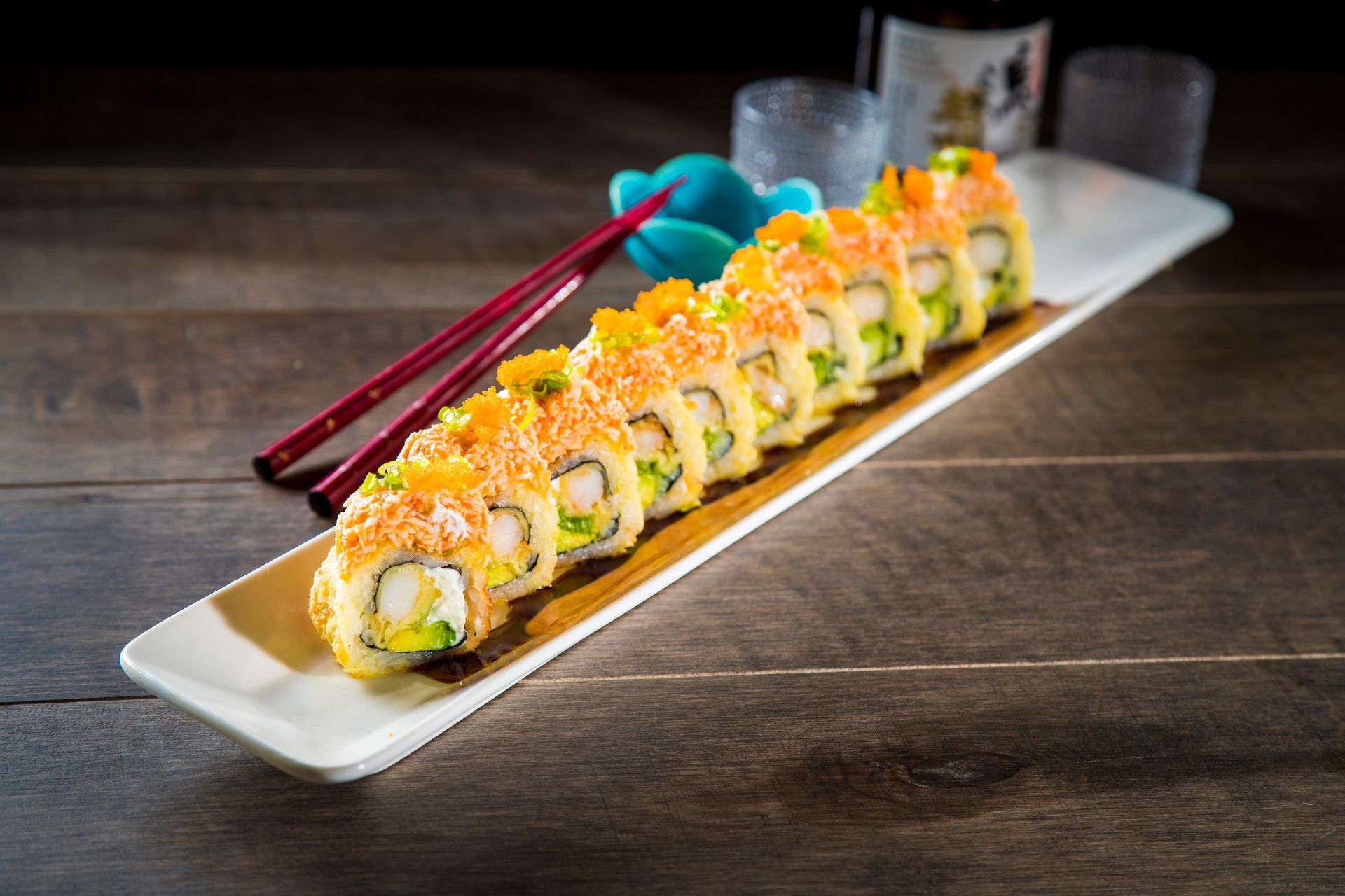 Sushi rolls topped with orange garnish on a long white plate, with chopsticks beside them on a dark table