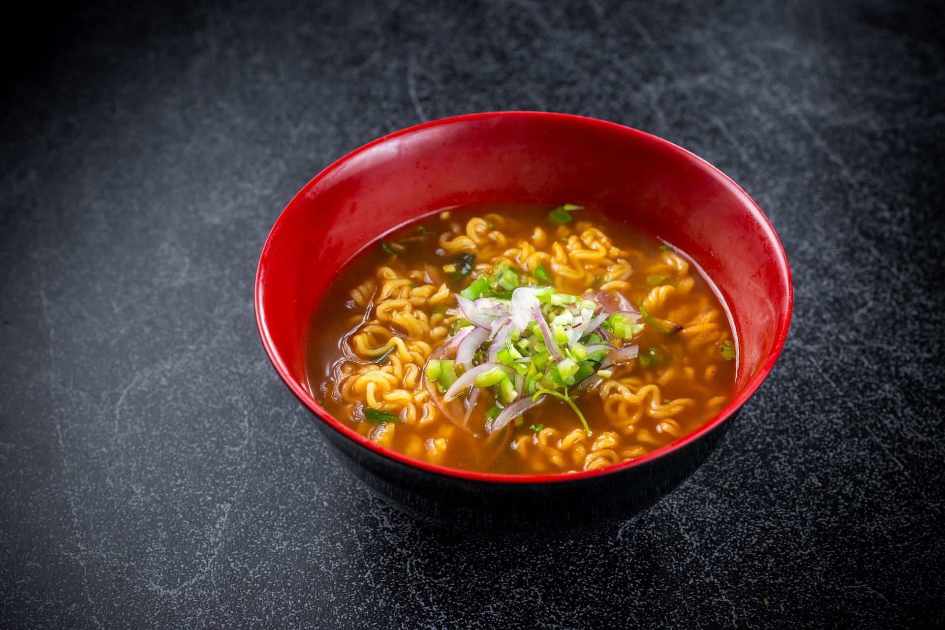 Red bowl of noodle soup with green garnish on a dark tabletop.