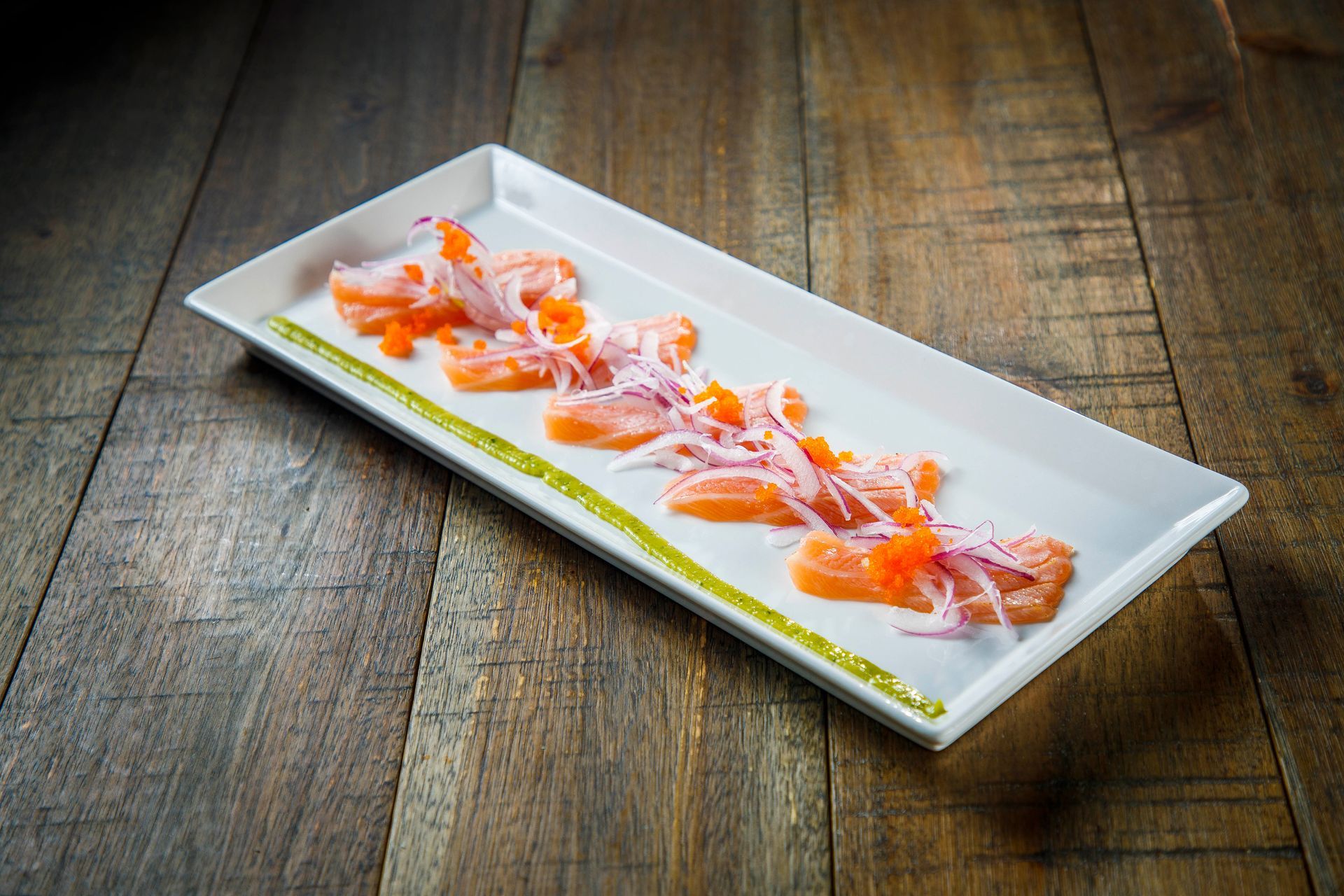 Slices of salmon sashimi on a white rectangular plate with green garnish on a wooden table