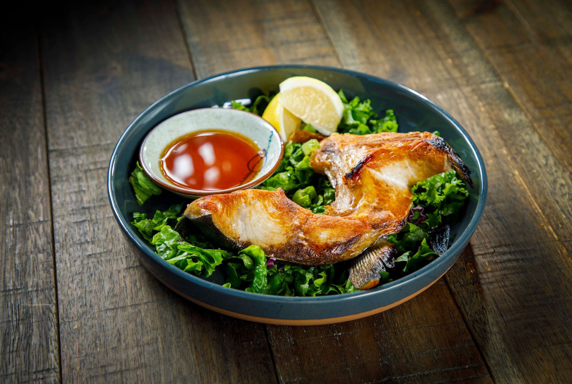 Grilled chicken leg on a dark bowl with green salad, lemon wedges, and dipping sauce on a wooden table