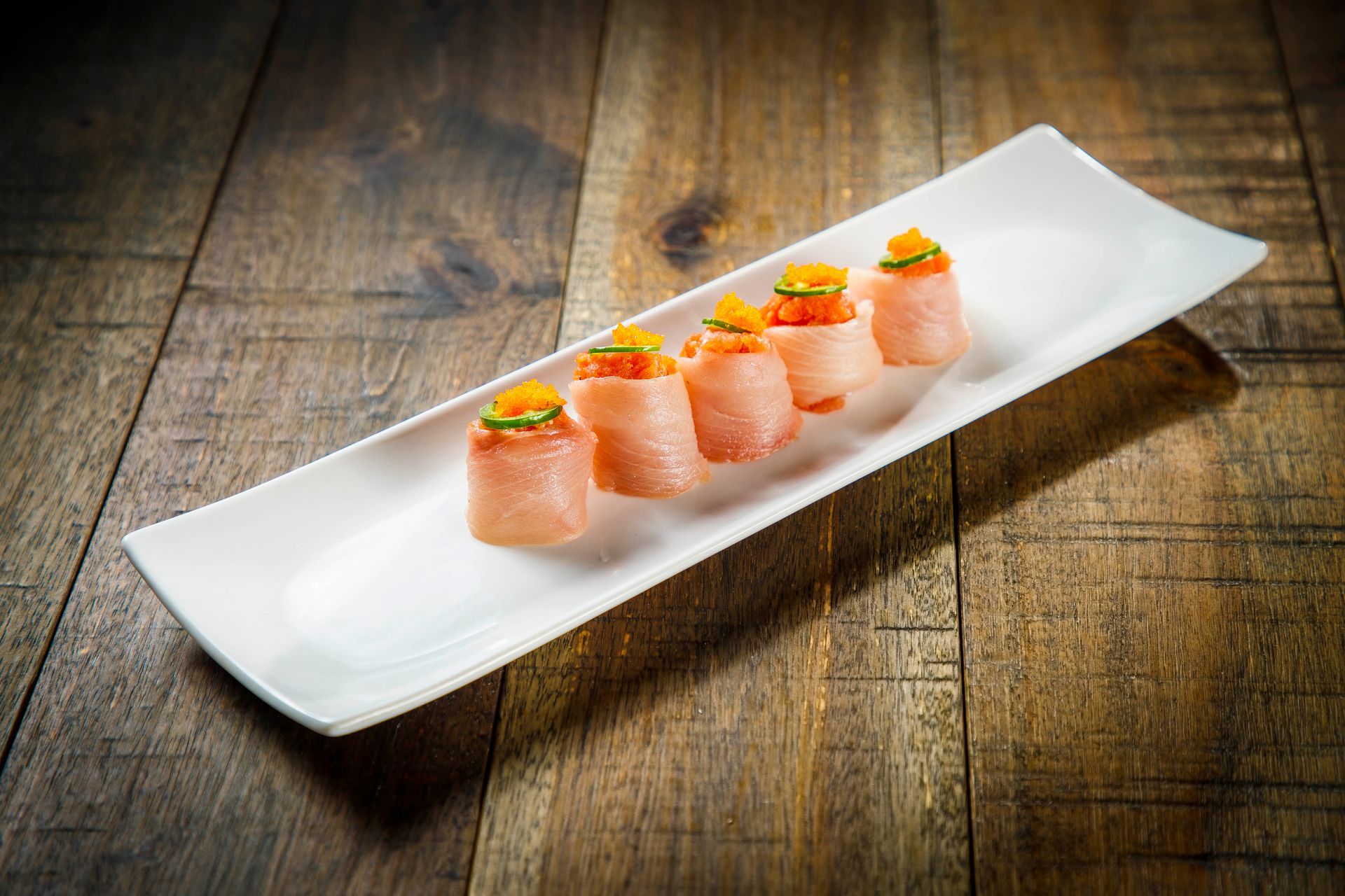 Five salmon sushi rolls topped with garnish on a long white plate on a wooden table