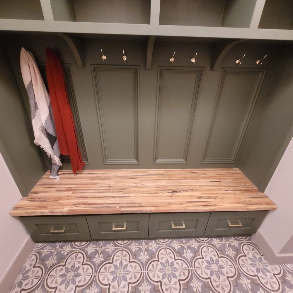 Green built-in mudroom with bench, drawers, hooks, and a wooden countertop.  Two scarves hang.  Ornate patterned floor tiles.