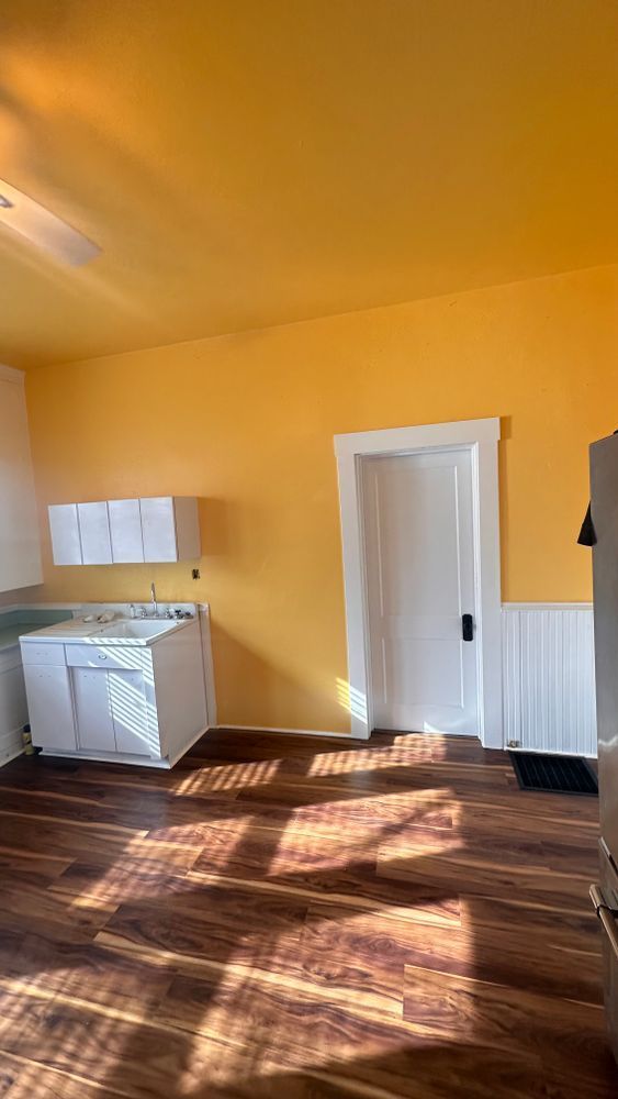 A kitchen with yellow walls and ceiling, white cabinets, and a wooden floor with sunlight.