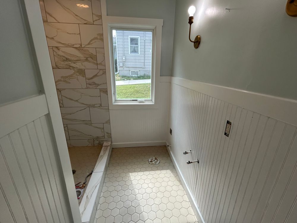 Bathroom with white wainscoting and hexagon tile floor. Shower with marble-like tiles, small window, and light blue walls.