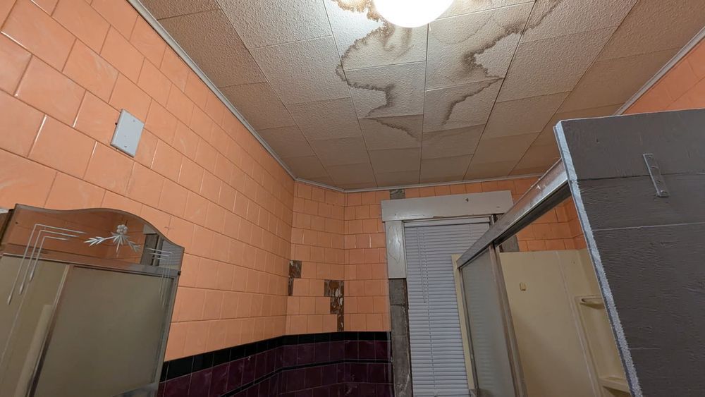 A public bathroom with orange tiled walls and a stained ceiling. A stall is open, and there's a mirror.