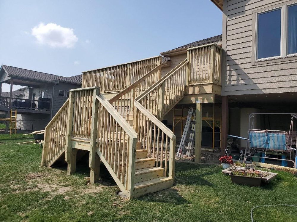 Wooden deck and stairs attached to a house, under a blue sky, with a lawn and other houses visible.
