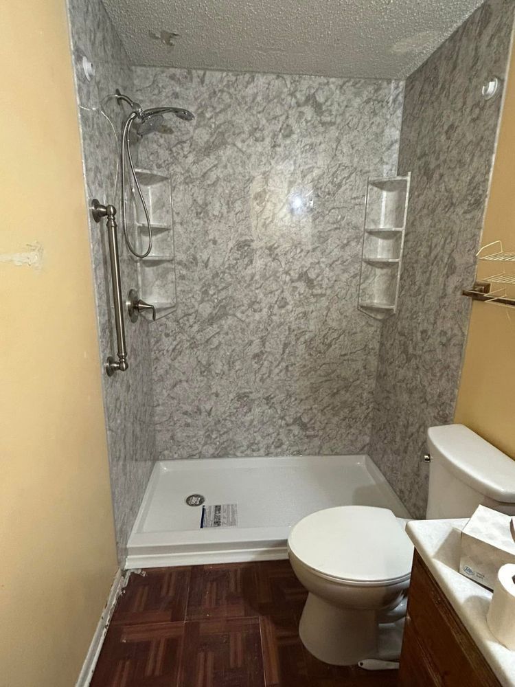 Shower with marbled gray walls, shelves, and chrome fixtures. Toilet on the right, brown floor.