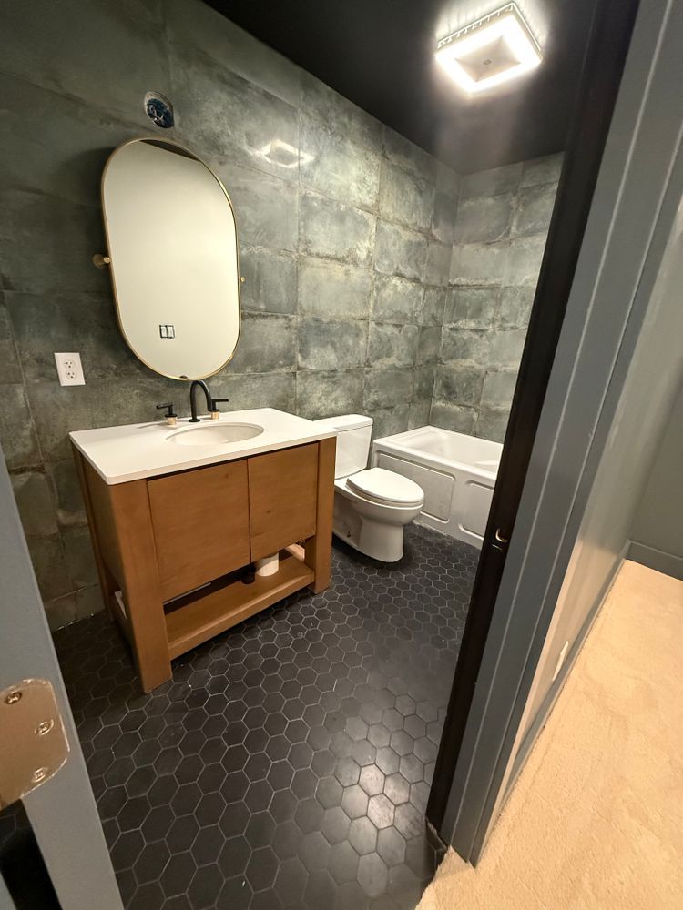 Bathroom with stone-like wall, wood vanity, oval mirror, black hexagon tile floor, toilet, and bathtub.