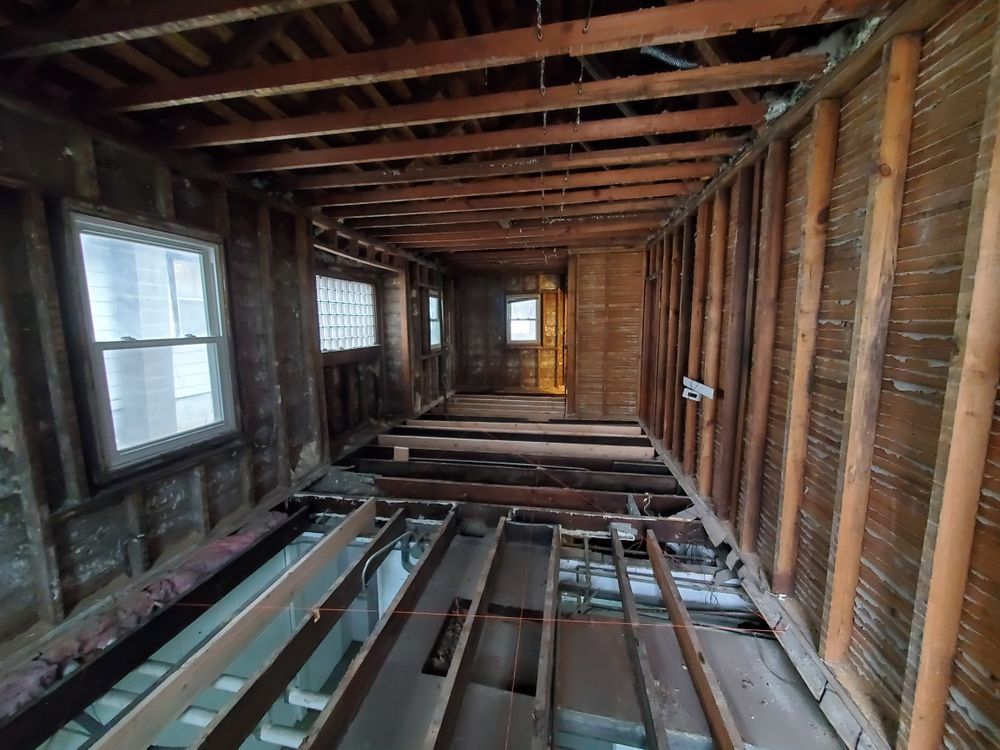 Interior room under renovation; exposed wood beams, studs, and floorboards. Windows are visible in the walls.