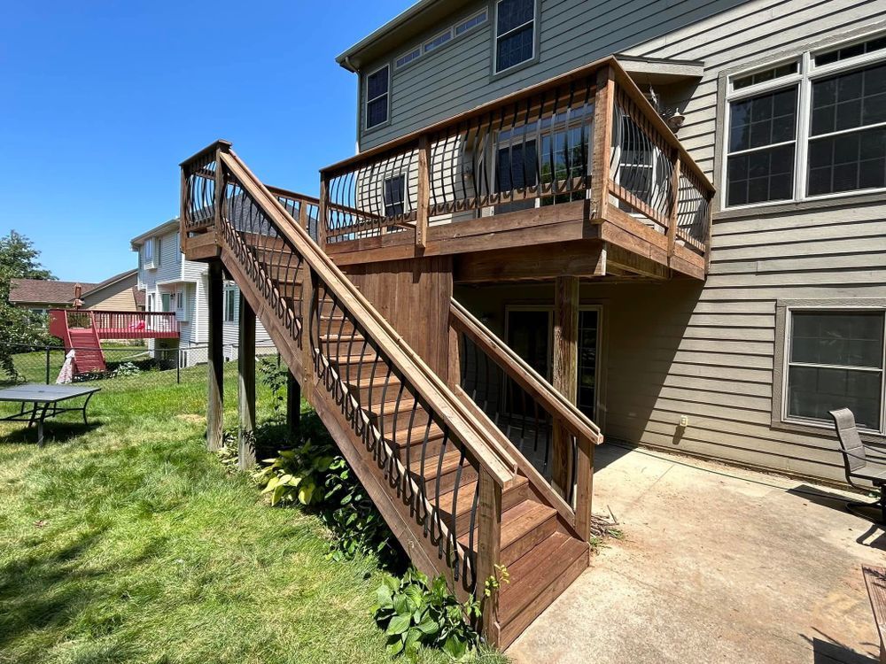 Wooden deck with stairs, attached to a two-story house. Brown stain, black spindles, concrete patio below.
