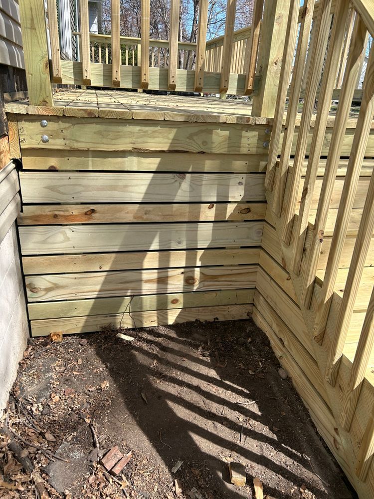 Wooden deck with horizontal siding, stairs, and handrails. Sunlight casts shadows on the ground.