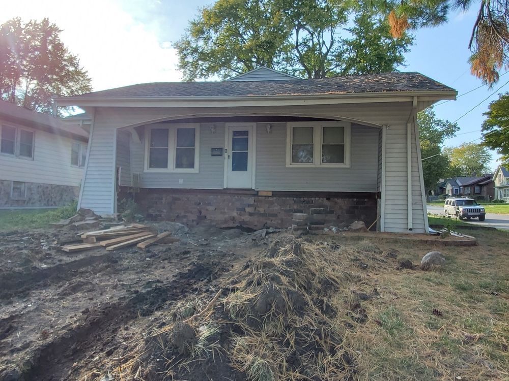 House under construction; light blue siding, brick foundation, porch. Dirt and trenches in front.