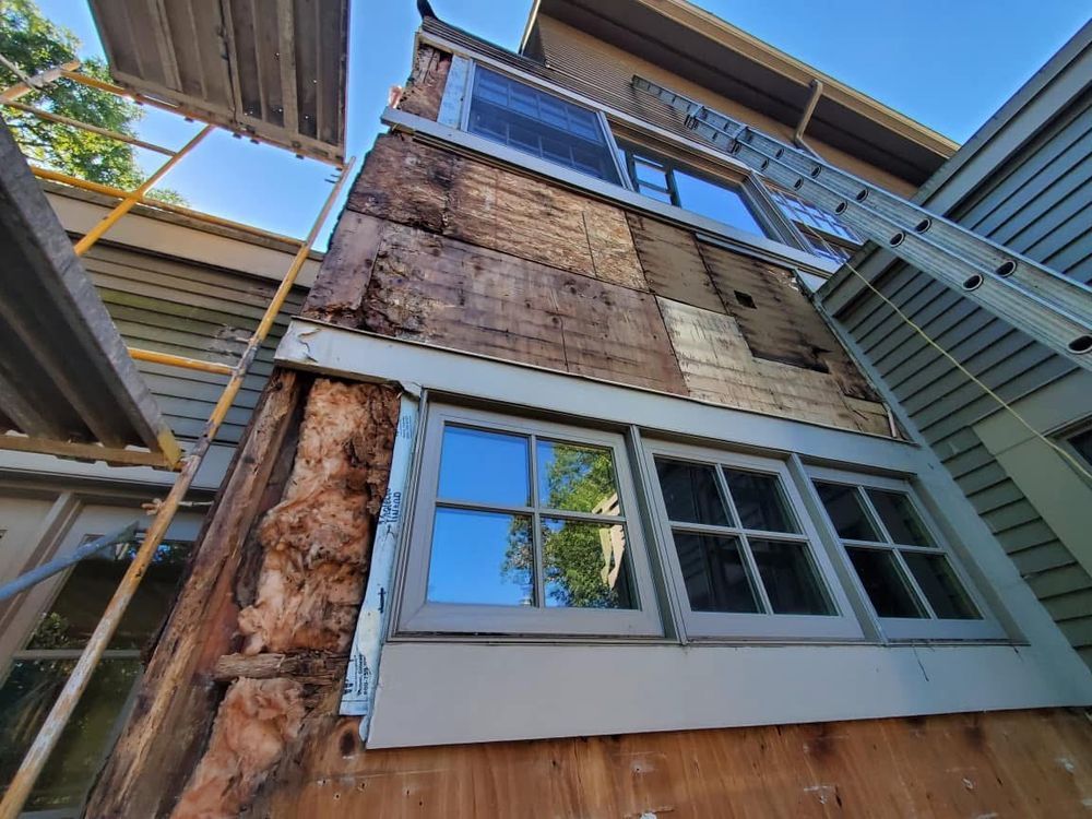 Exterior house wall under renovation, showing wood damage, exposed frame, and windows. A ladder and scaffolding are present.