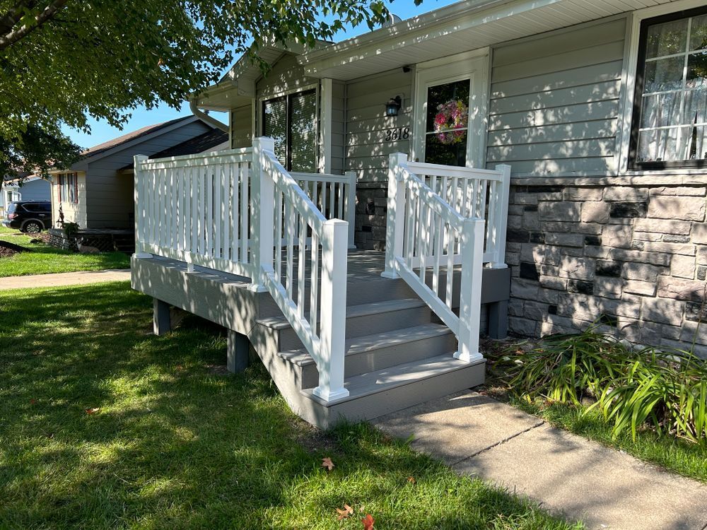 White railing surrounds a gray porch with steps leading to a house. Green grass and landscaping.