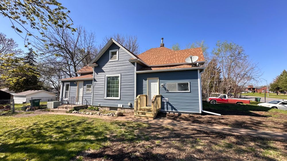 Two-story blue house with brown roof and yard under a blue sky. Red truck visible in the background.