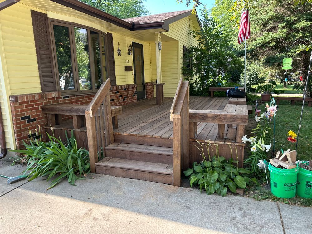 Yellow house with wooden porch, steps, and American flag. Garden with green buckets on the right.