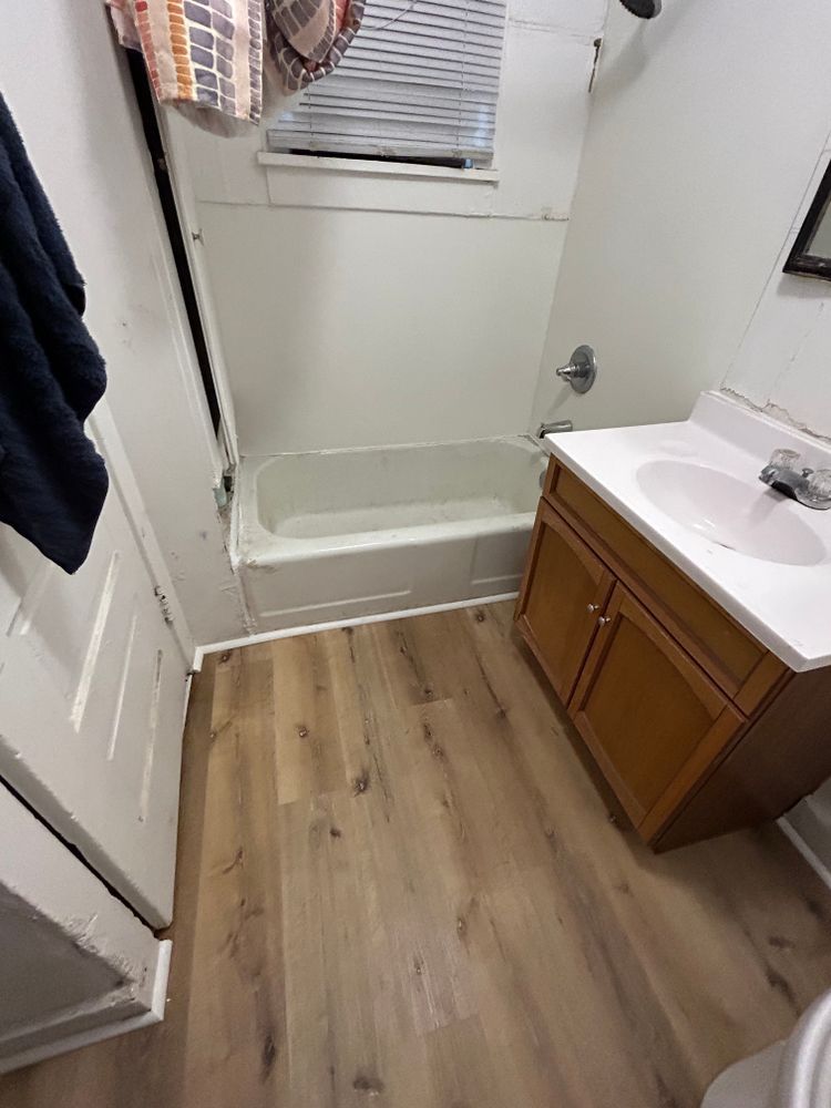 Small bathroom with a tub, vanity, and wood-look flooring. White walls, a window with blinds, and a partially visible door.