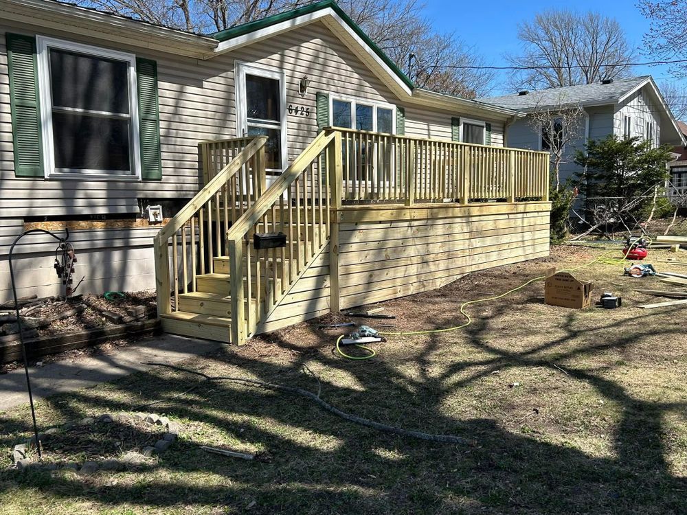 Wooden deck with steps built onto a beige house. Green shutters and trim. Sunny outdoors.