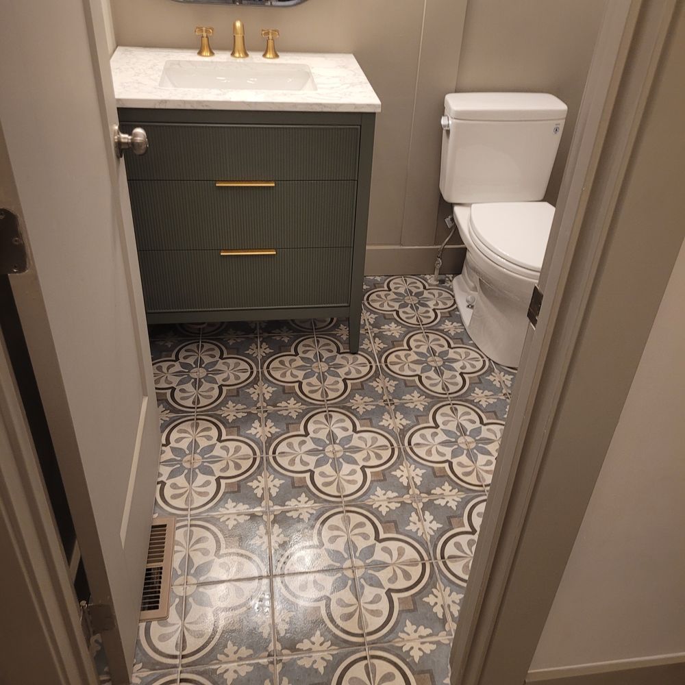 Small bathroom with ornate patterned floor tiles, green cabinet, white toilet, and gold fixtures.