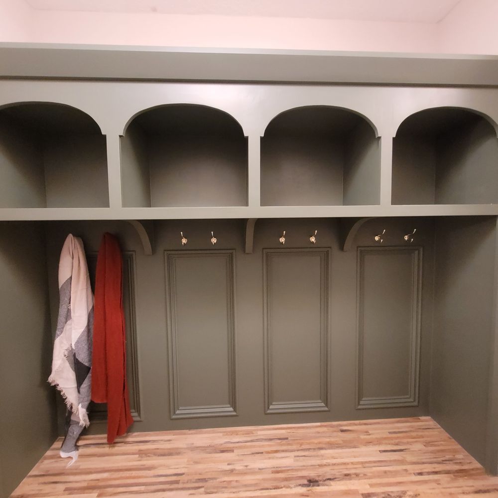 Built-in olive green storage unit with arched shelves, hooks, and paneling. Scarves hang; wooden floor.