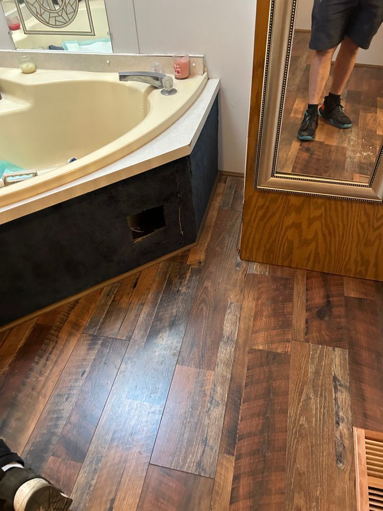 Bathroom interior: bathtub with black front, wood-look floor, and partial mirror reflection of a person.