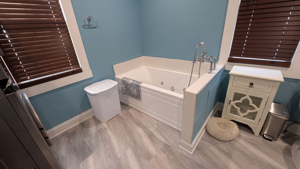 Bathroom with built-in tub, blue walls, gray wood-look floor, wooden blinds, and a white side table.