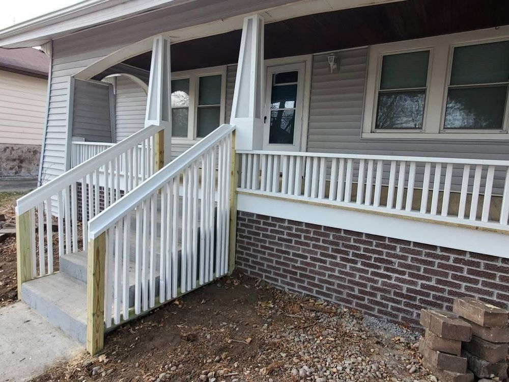Exterior of house with new white porch railing and steps, gray siding, and brick foundation.