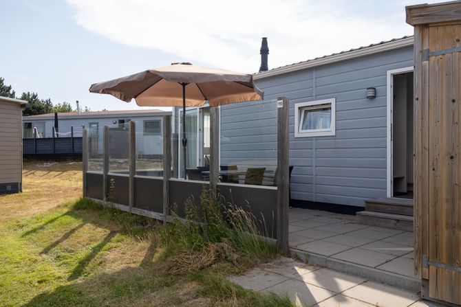 Terras met beige parasol en glazen balustrade naast een lichtblauw huis op een zonnige dag.