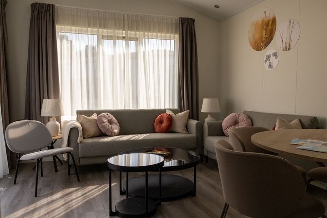 Gezellige woonkamer met grijze bank, ronde salontafel, fauteuils, eettafel en groot raam met gordijnen.
