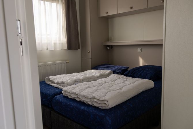Kleine slaapkamer met een netjes opgemaakt tweepersoonsbed, blauw beddengoed en een raam met planken boven het bed.