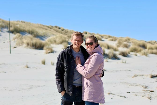Twee mensen staan ​​op een zandstrand met duinen en lachen naar de camera.