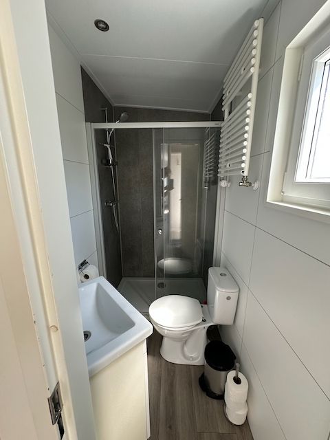 Smalle witte badkamer met douche, toilet, wastafel en raam aan de rechterkant.