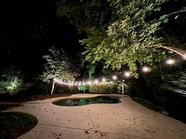 A swimming pool is lit up at night with a string of lights hanging from trees.